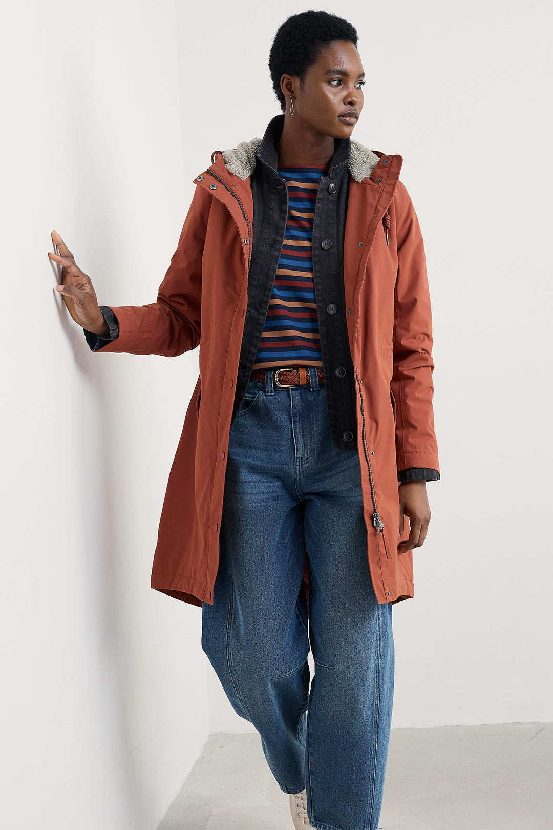 Person wearing a long rust-colored coat over a striped shirt and jeans on a white background