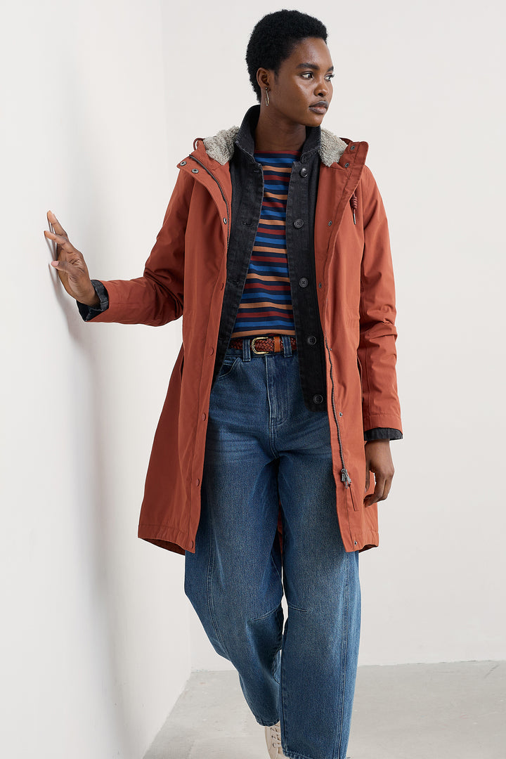 Person wearing a long rust-colored coat over a striped shirt and jeans on a white background