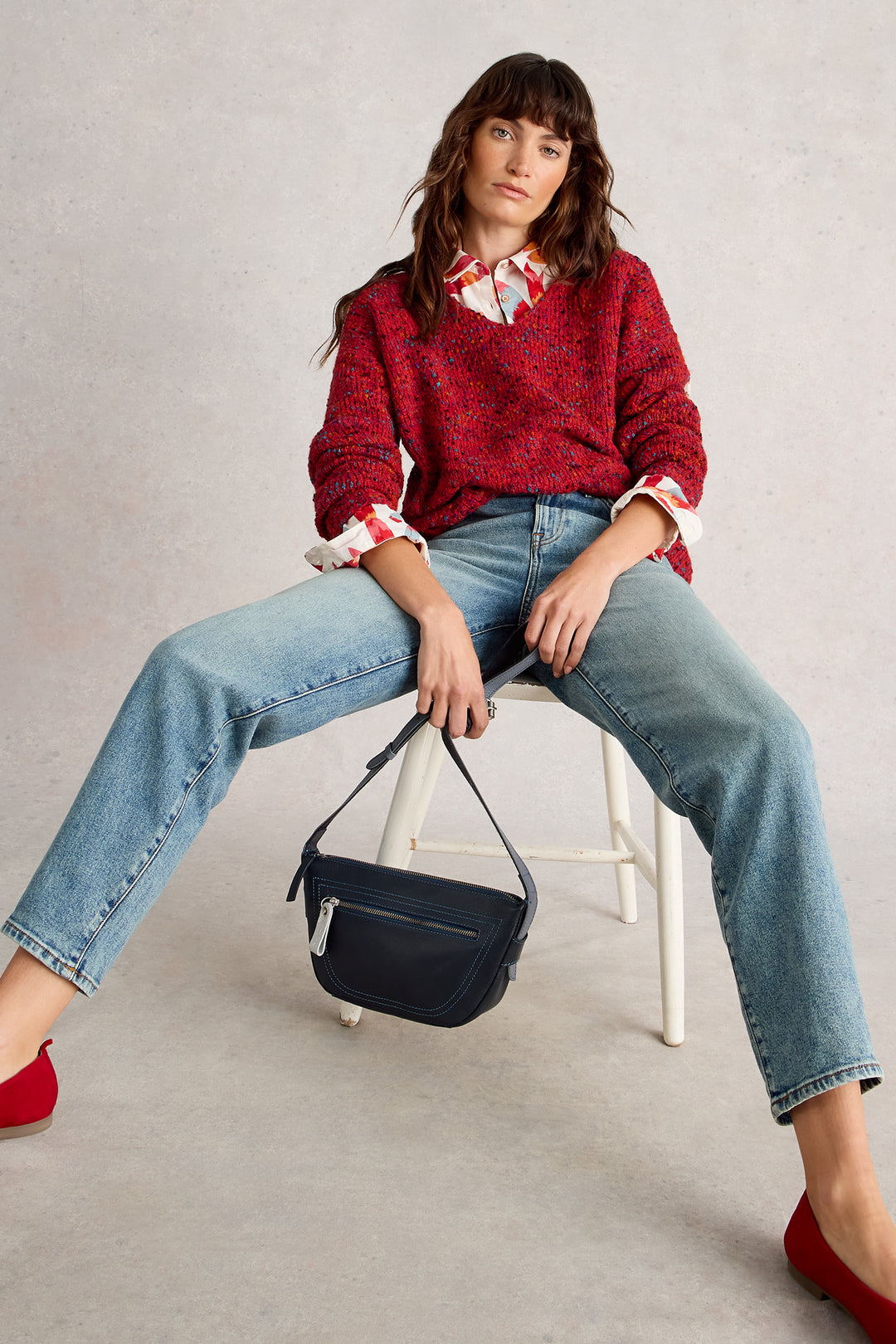 Woman sitting on a stool holding a black handbag against a plain background