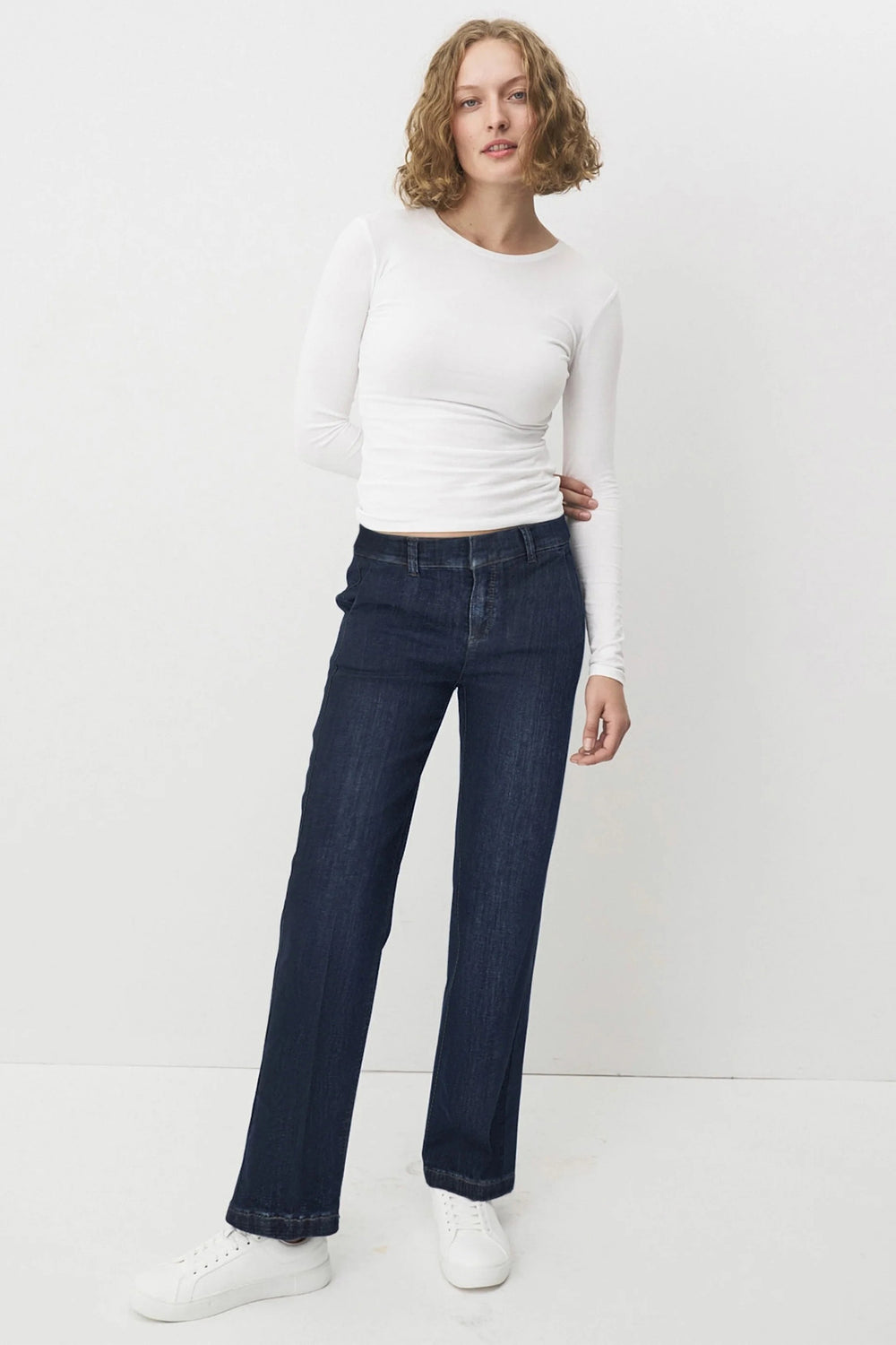 Woman wearing a white long-sleeve top and dark blue jeans on a white background