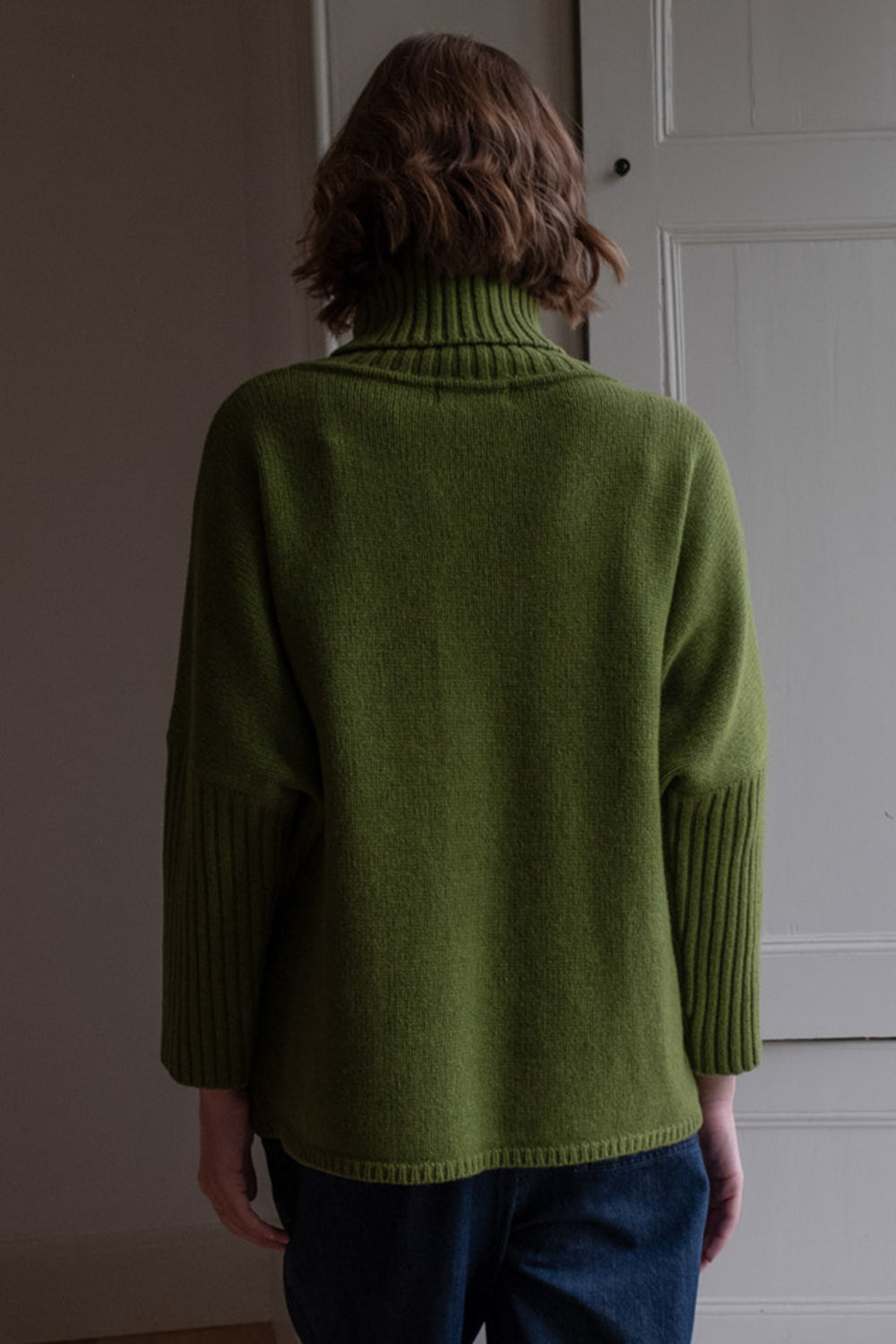 Person wearing a green sweater standing in front of a white door.
