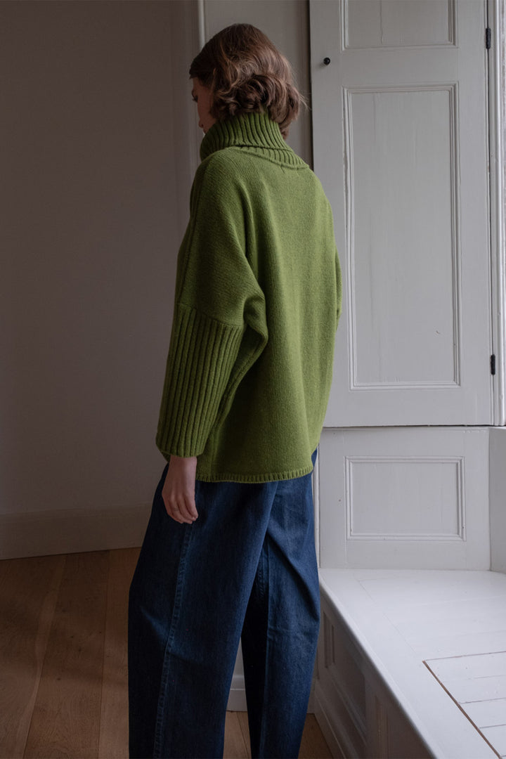 Person wearing a green sweater and blue pants standing in a room with white walls and a door.