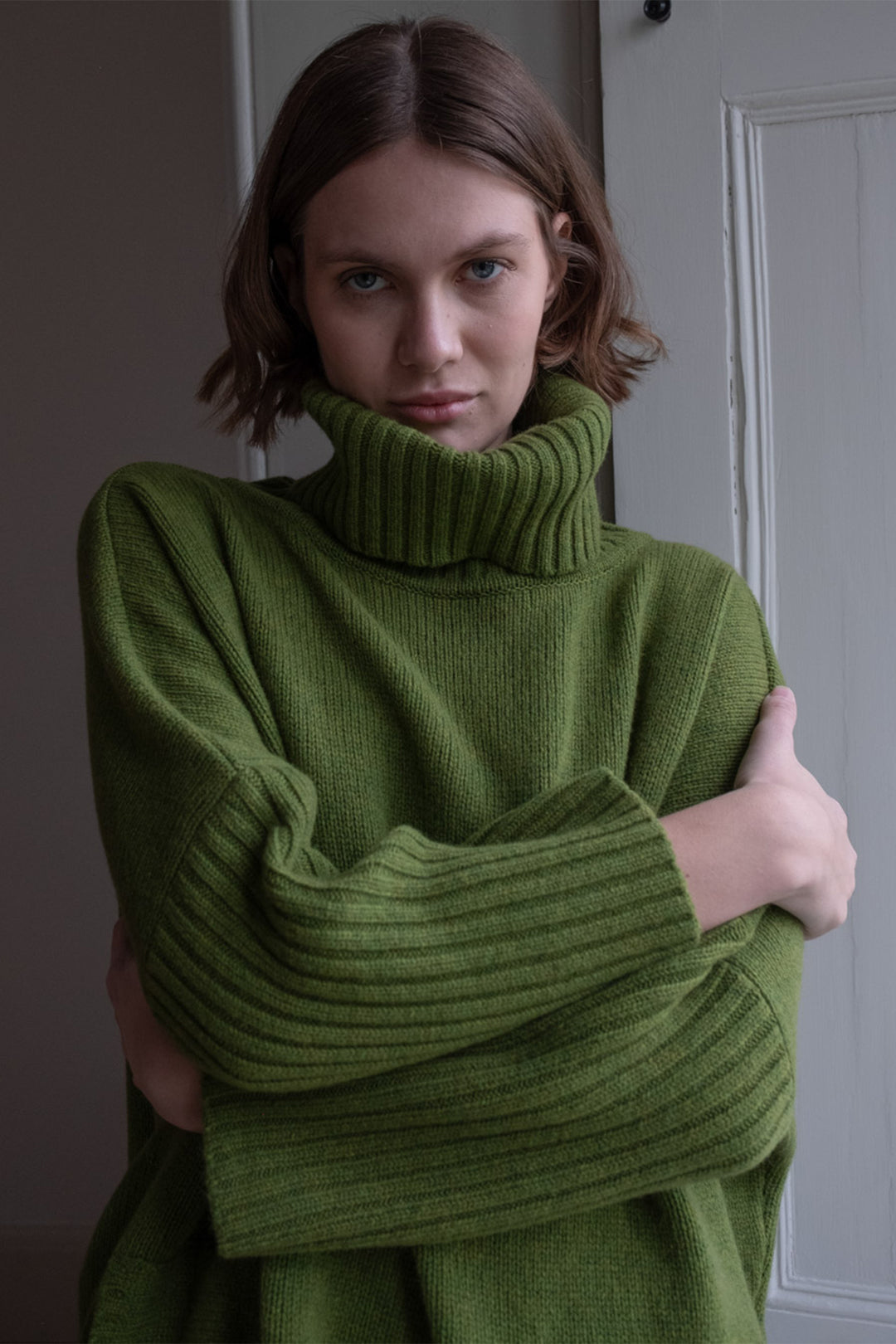 Woman wearing a green sweater with her arms crossed in a room.