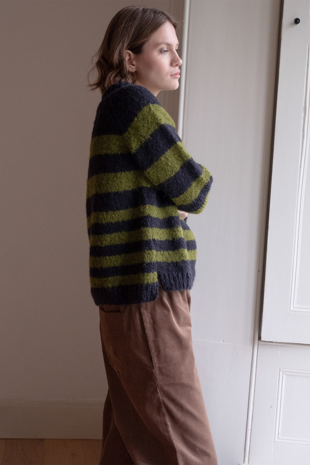 Woman wearing a striped sweater and brown pants standing against a plain wall.
