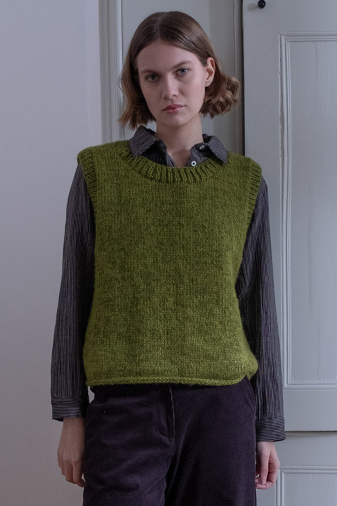 Person wearing a green knitted vest over a dark shirt and pants in a neutral setting.