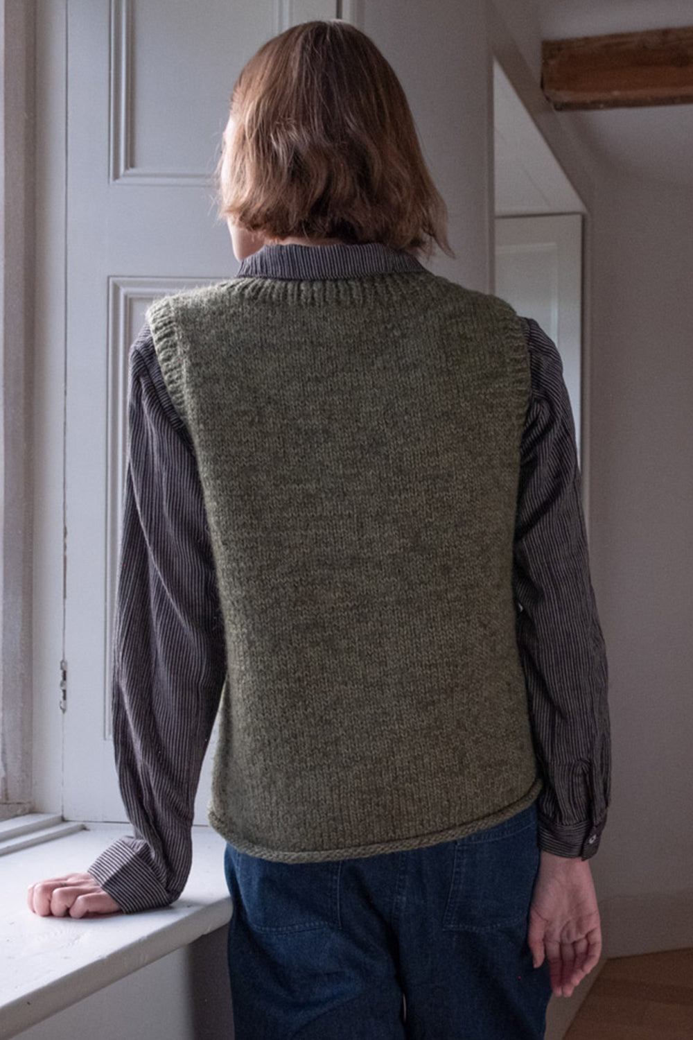 Person wearing a green knitted vest in a room with a window.