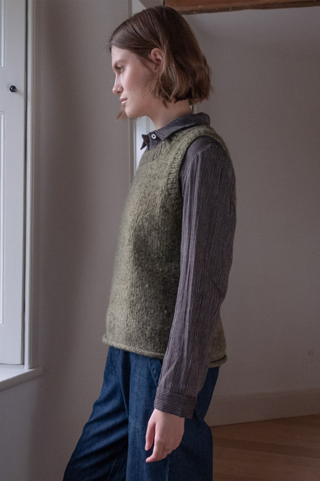 Person wearing a green knitted vest over a long-sleeve shirt and blue pants in a room.