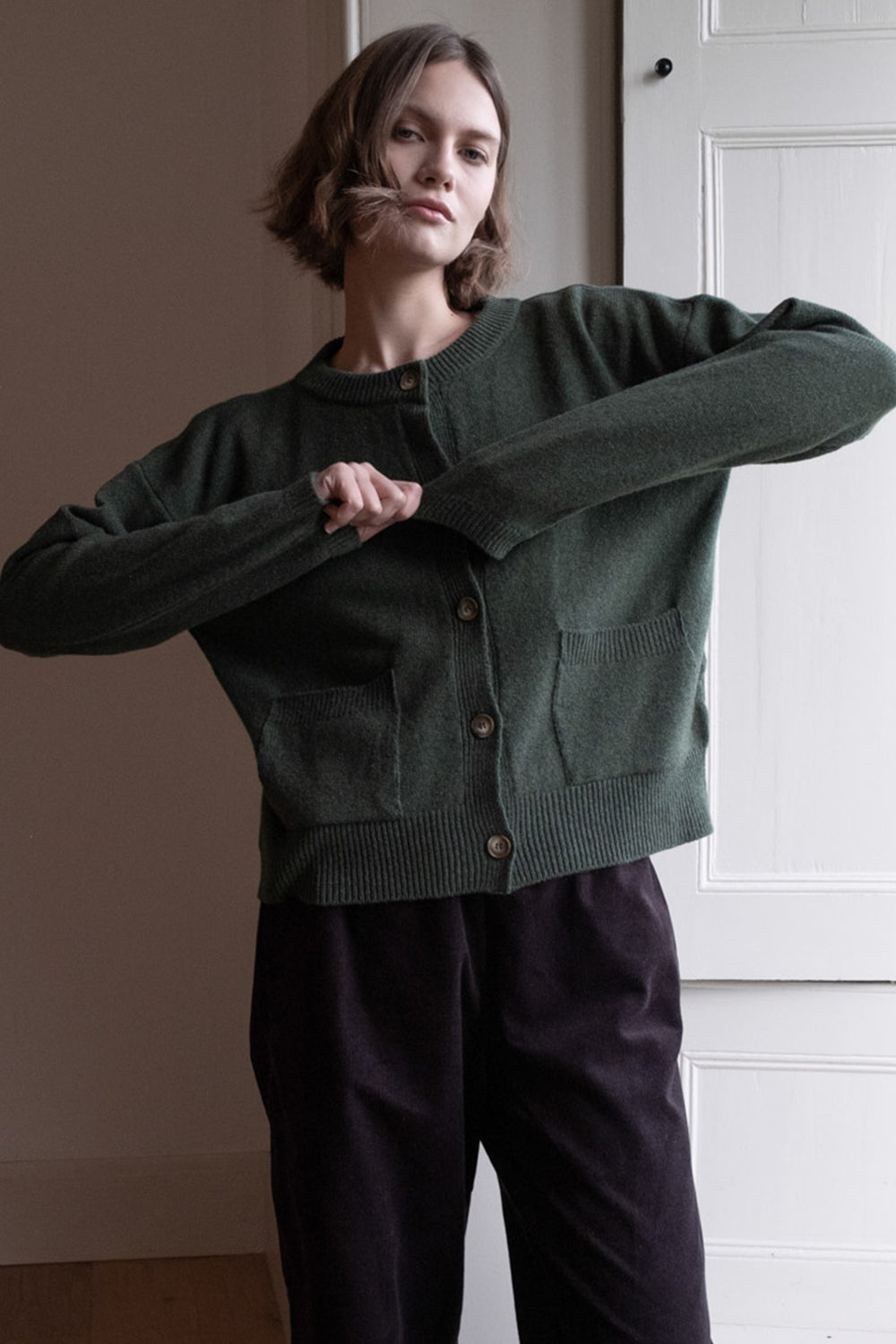 Person wearing a green cardigan and dark pants in a minimal indoor setting