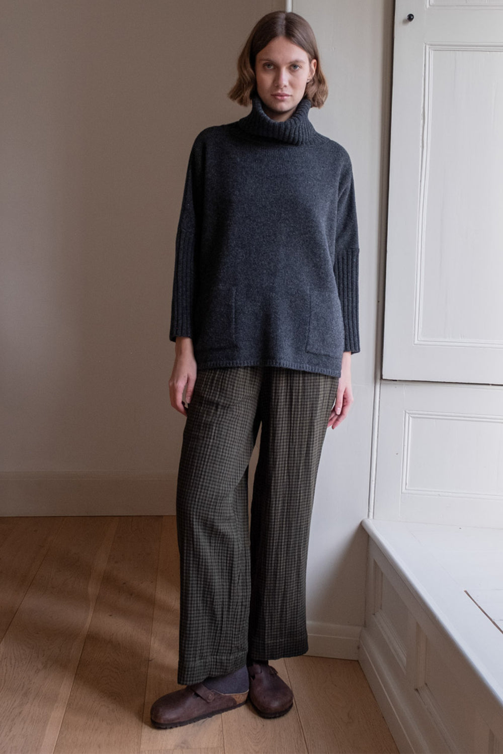Person wearing a dark sweater and plaid pants standing in a room with wooden floor and white walls.