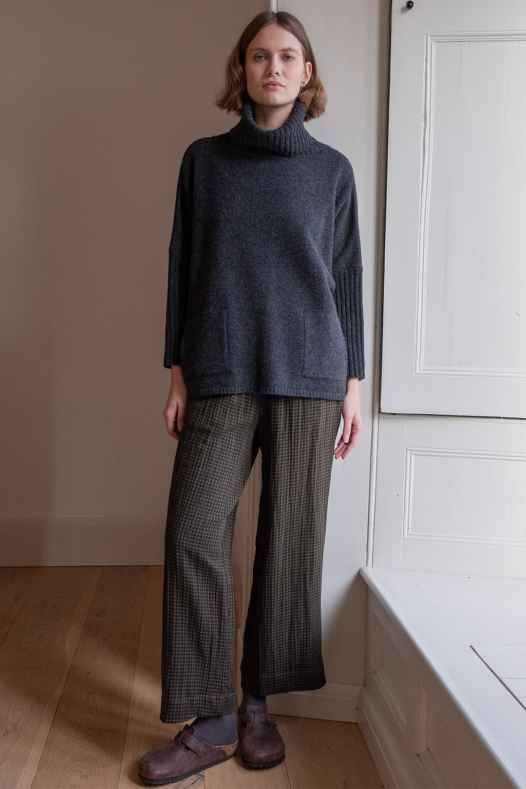 Person wearing a dark gray turtleneck sweater and plaid pants standing in a room with a neutral color palette.