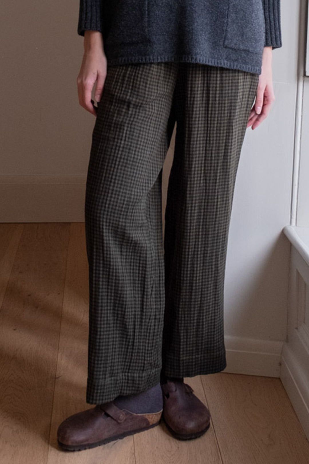 Person wearing plaid pants and a dark sweater in a room with wooden floor and white walls.