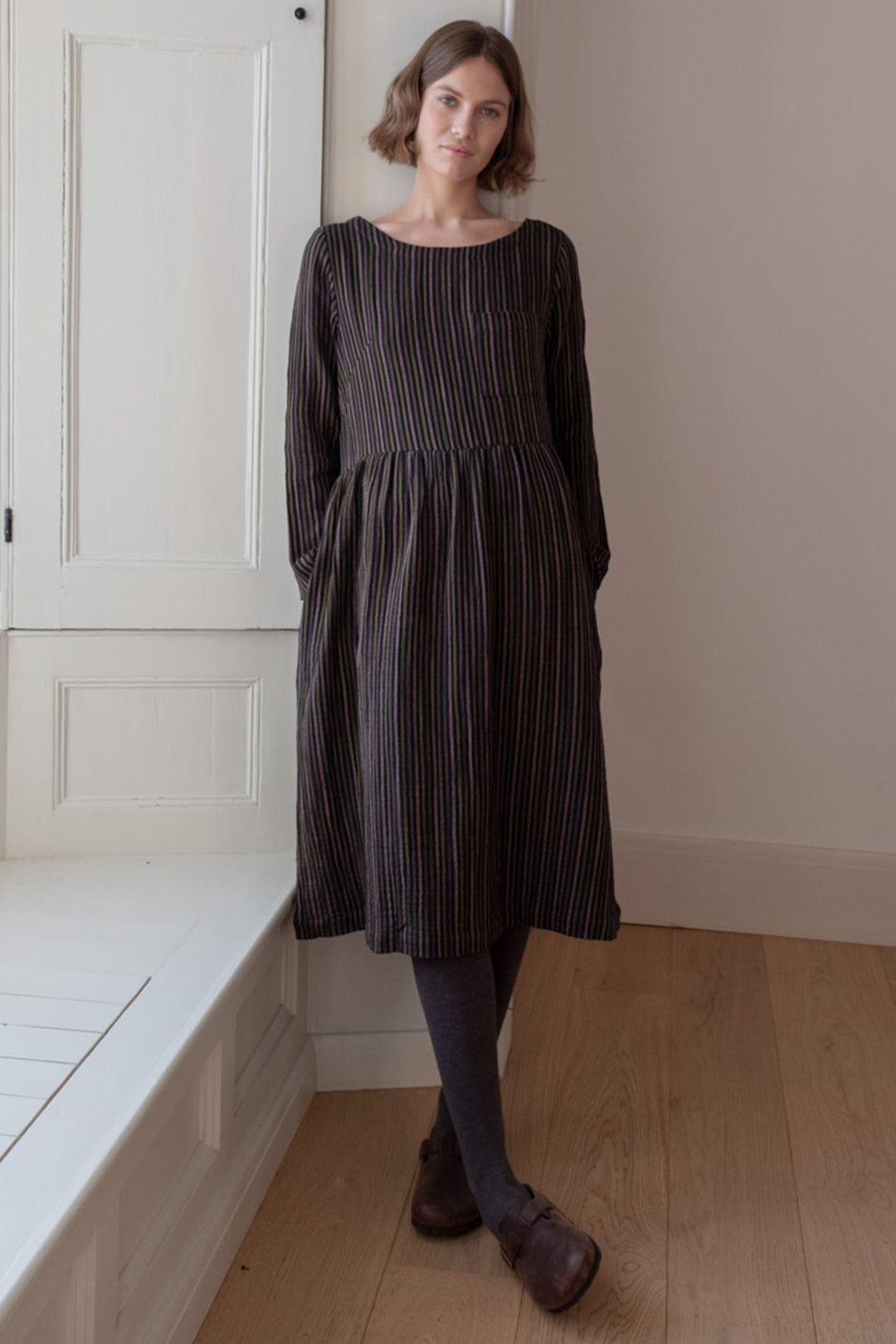Woman wearing a dark striped dress standing in a room with wooden flooring and white walls.