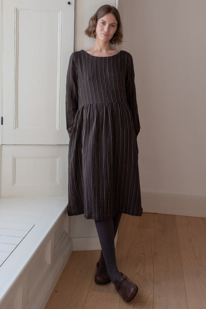 Woman wearing a dark striped dress standing in a room with wooden flooring and white walls.
