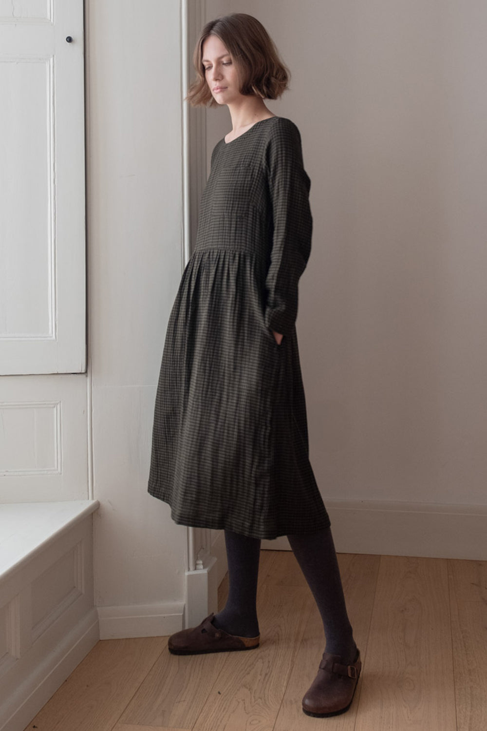 Woman wearing a dark green dress standing in a room with white walls and wooden floor.