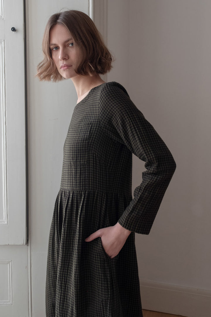 Woman wearing a dark green checkered dress standing against a plain wall.