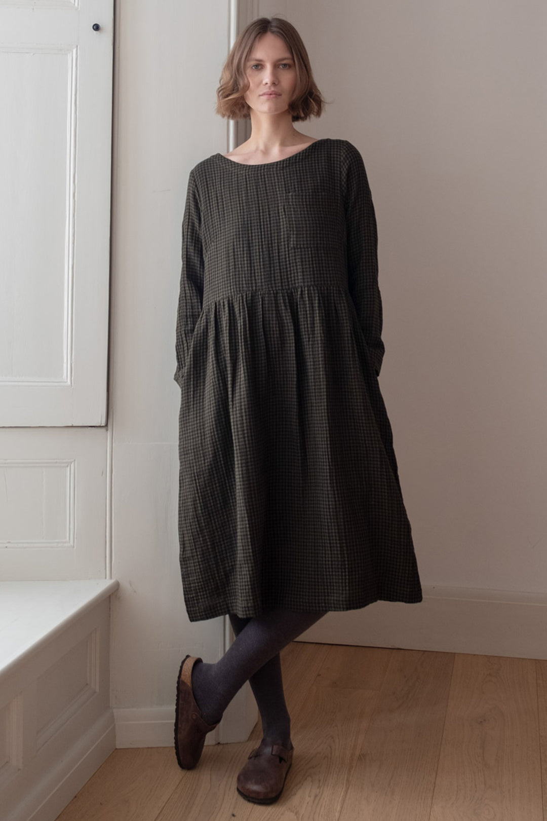 Woman wearing a dark green dress standing in a room with white walls and wooden floor.