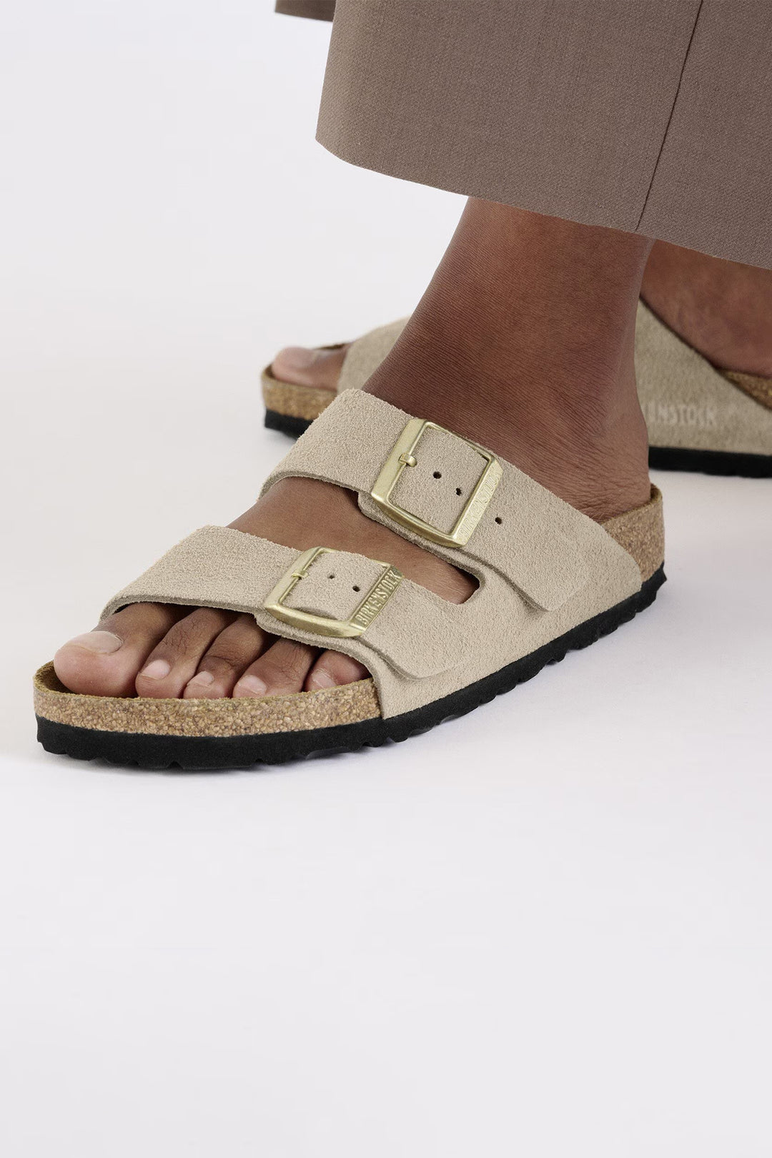 Beige sandals with a gold buckle worn by a person on a white background