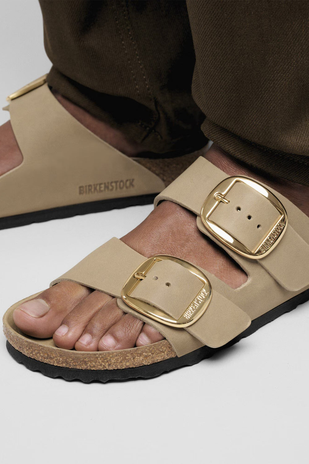 Beige sandals with gold buckles worn by a person on a white background