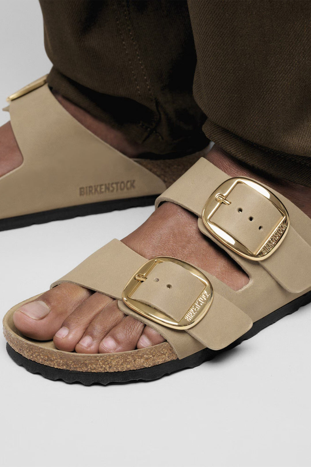 Beige sandals with gold buckles worn by a person on a white background