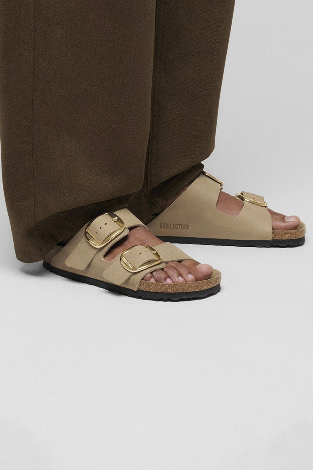 Beige sandals with gold buckles worn by a person in brown pants on a light gray background