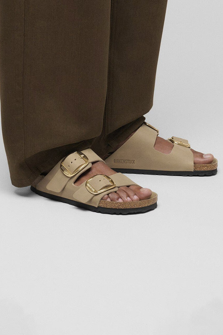 Beige sandals with gold buckles worn by a person in brown pants on a light gray background