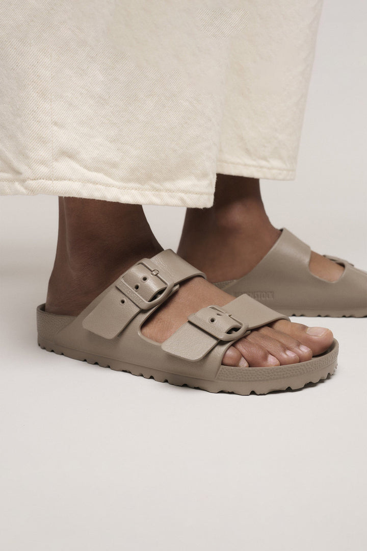 Beige sandals worn with a white skirt on a neutral background