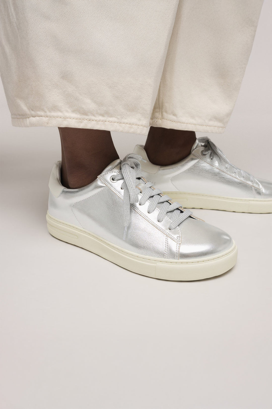 Silver sneakers with white soles worn with light-colored pants on a plain background