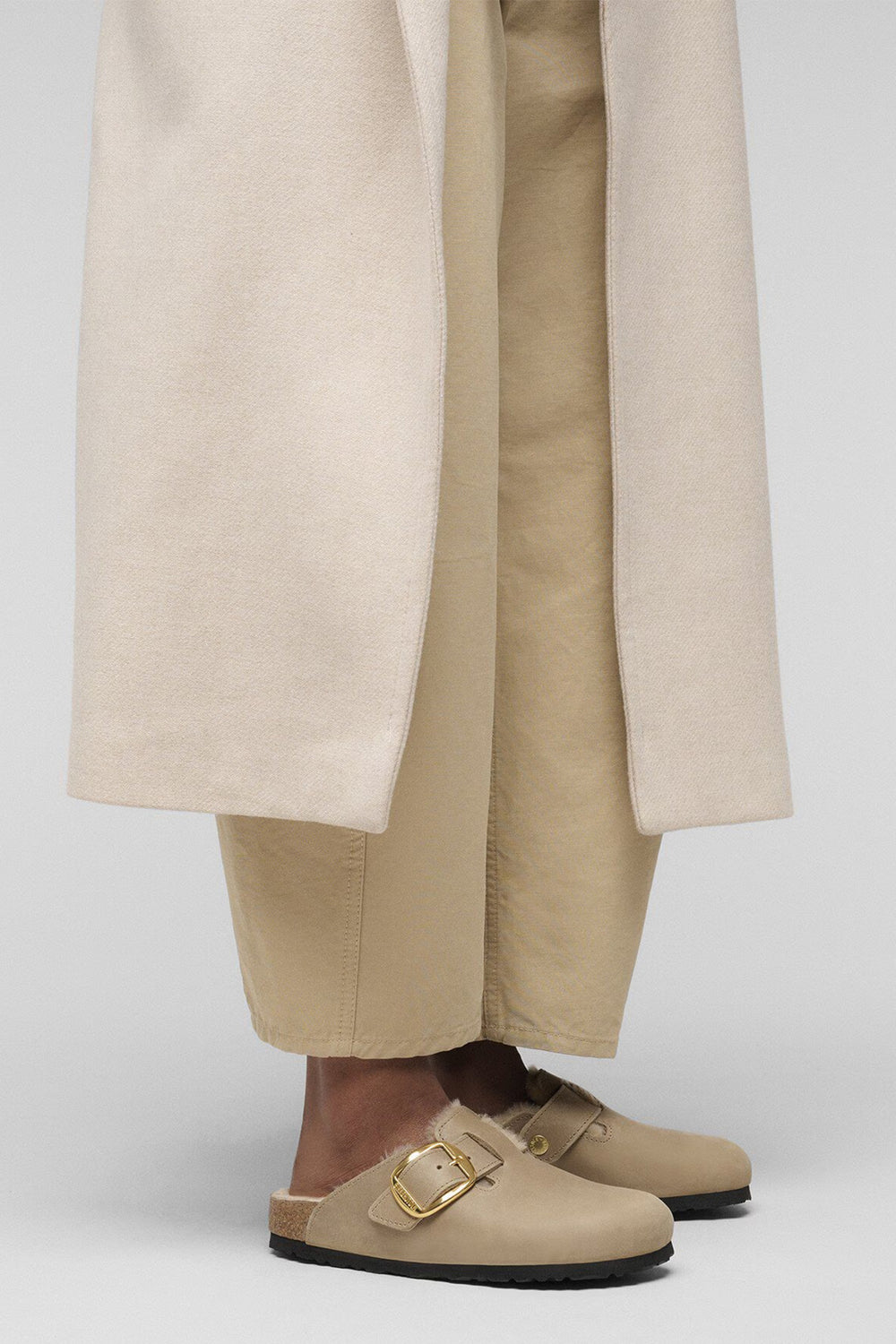 Person wearing beige shoes with a gold buckle, paired with beige pants on a white background