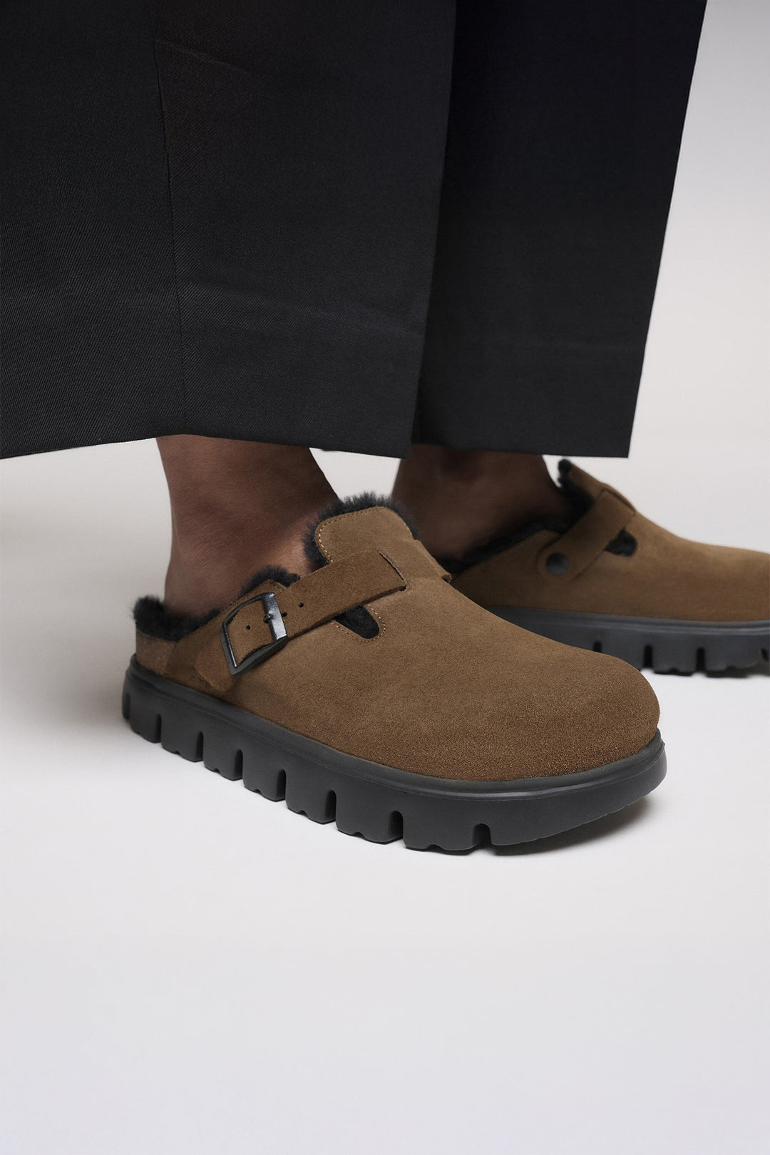 Brown suede shoes with black soles worn with black pants on a light background