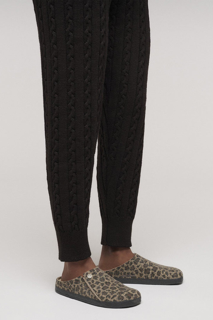 Person wearing black cable-knit pants and leopard print slippers on a white background