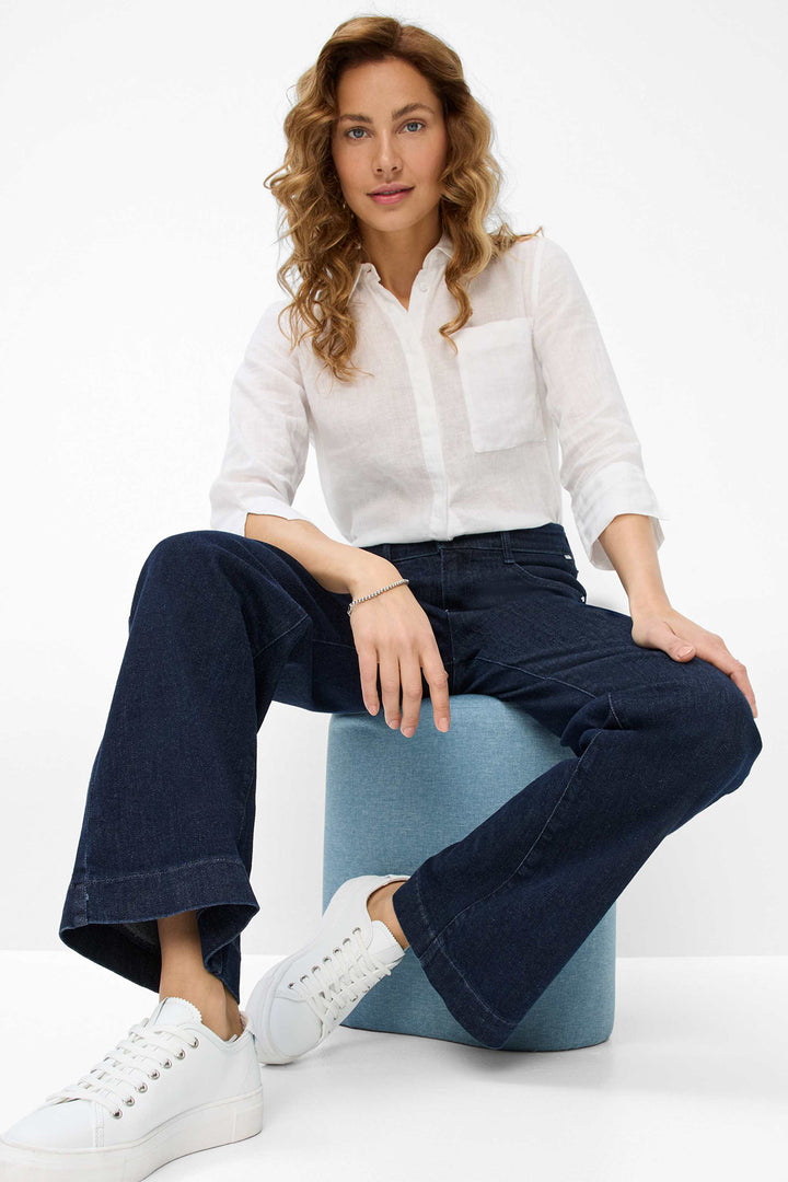 Woman sitting on a blue stool wearing a white shirt and dark blue pants with white sneakers.