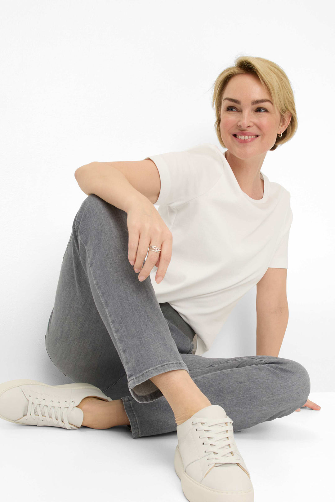 Woman sitting on a white background wearing a white top, gray pants, and white sneakers.