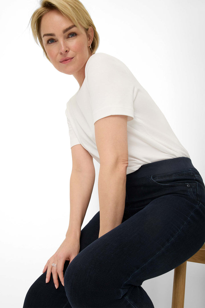 Woman wearing a white t-shirt and dark jeans sitting on a stool against a white background