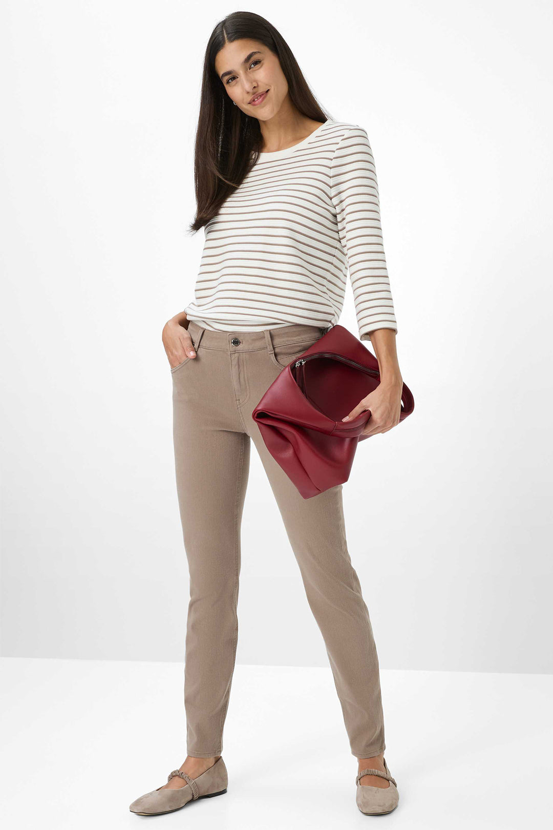 Woman wearing a striped shirt and beige pants holding a red clutch against a white background