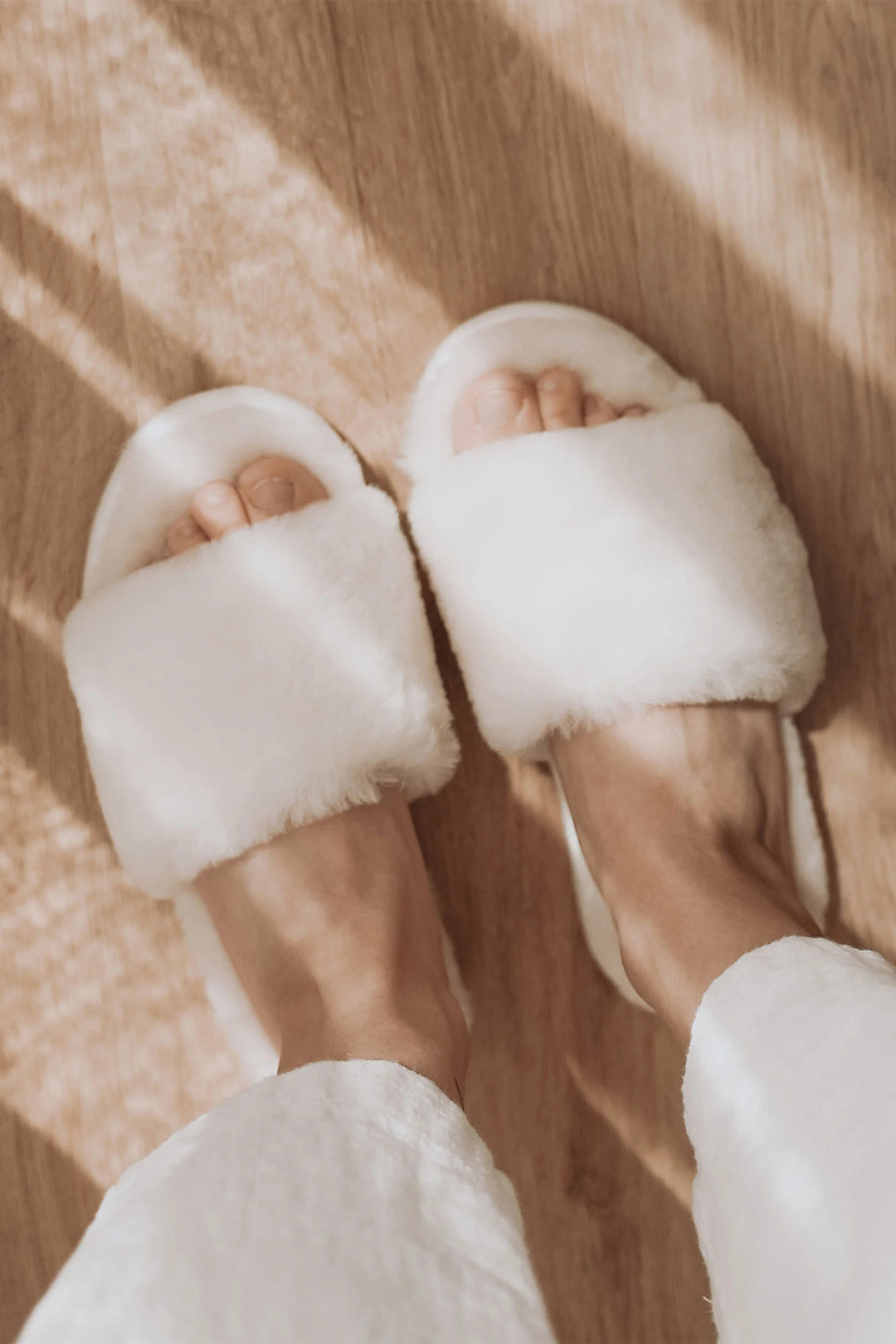 White fluffy slippers worn on a wooden floor with sunlight casting shadows.