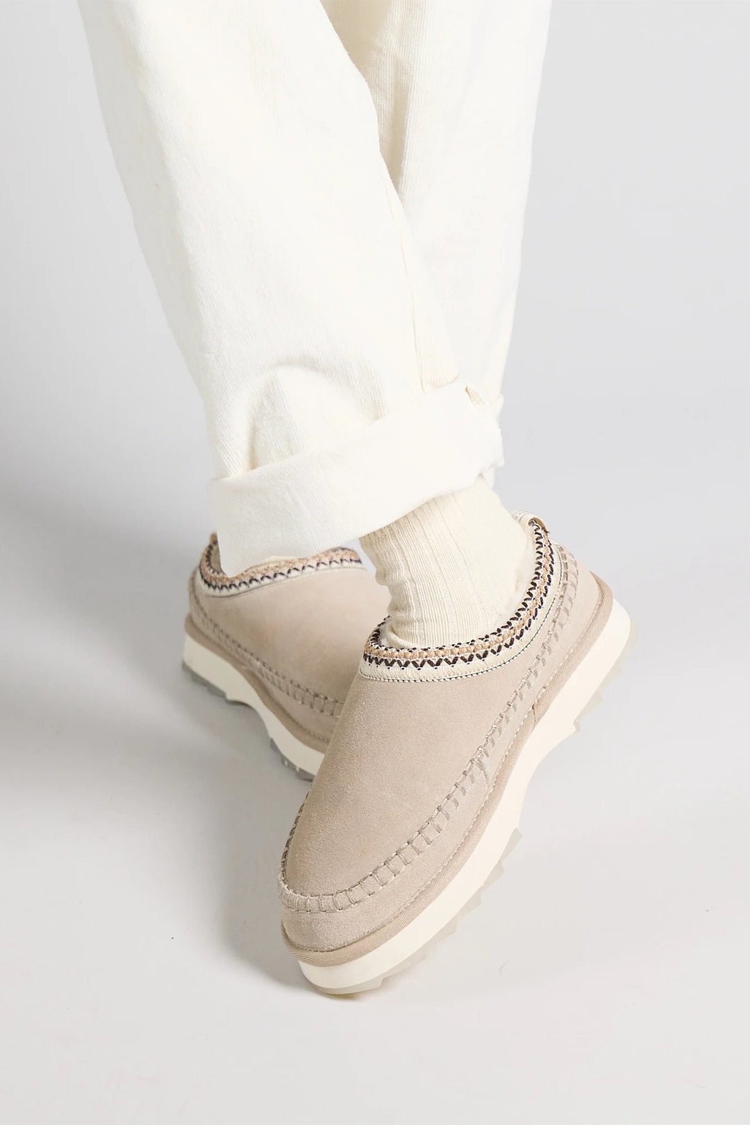 Beige slip-on shoes worn with white pants on a light gray background