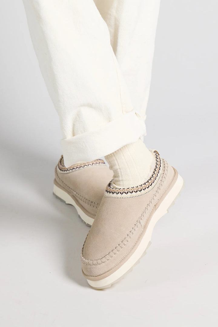 Beige slip-on shoes worn with white pants on a light gray background