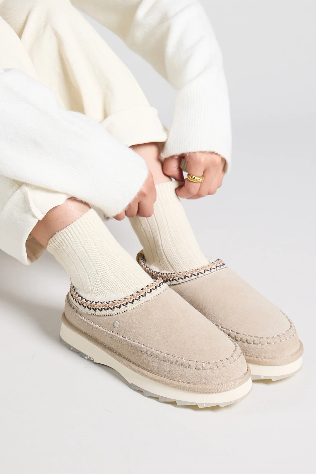 Person wearing beige moccasin-style shoes with decorative trim on a light gray background