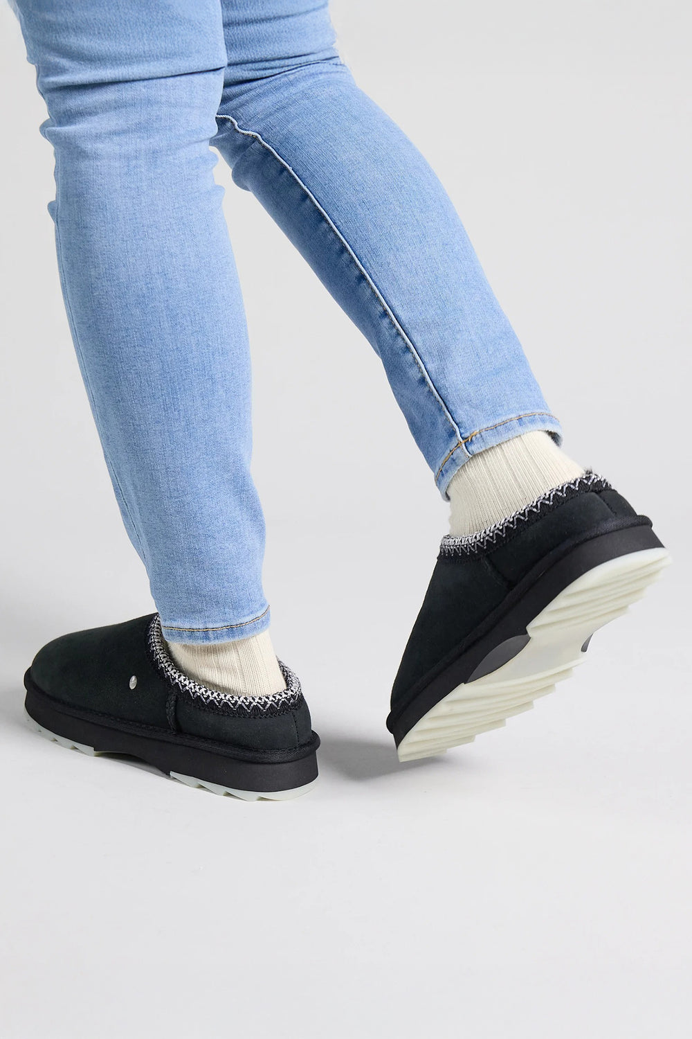 Person wearing black shoes with white soles and light blue jeans on a gray background