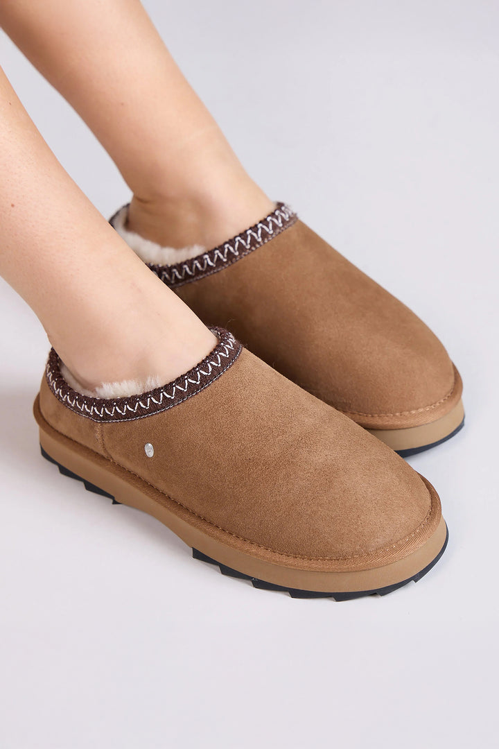 Brown suede slippers with decorative trim worn by a person on a light gray background