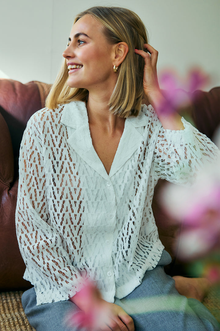 Woman wearing a white lace top sitting on a couch with blurred flowers in the foreground