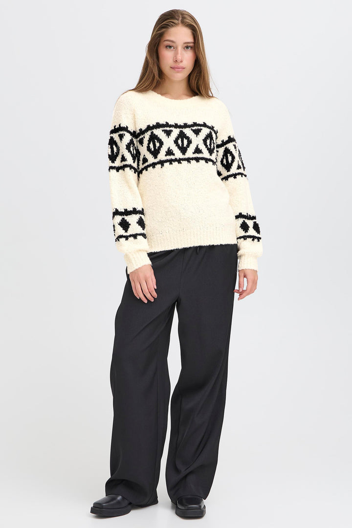 Woman wearing a cream sweater with black geometric patterns and dark pants on a white background