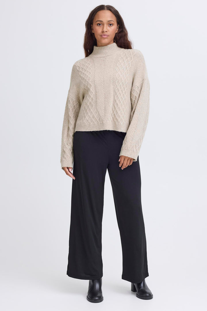 Woman wearing a beige cable knit sweater and black pants on a white background