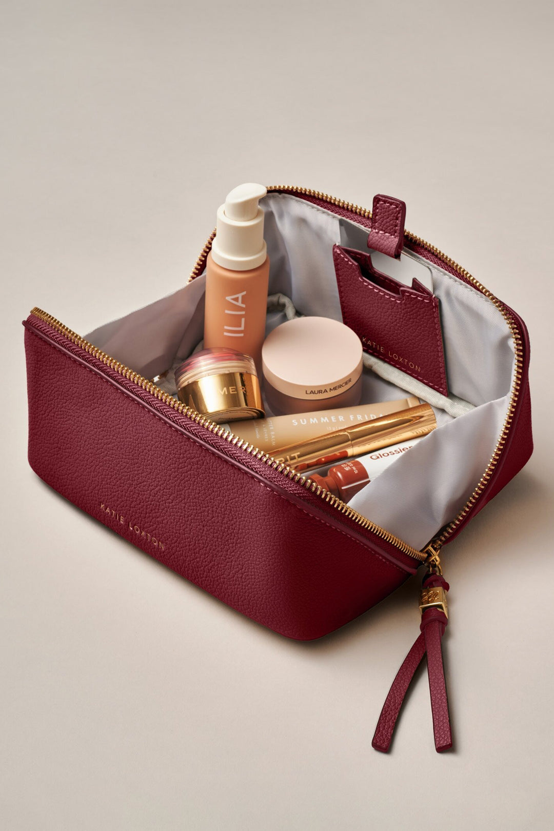 Open red cosmetic bag with ILIA products on a beige background