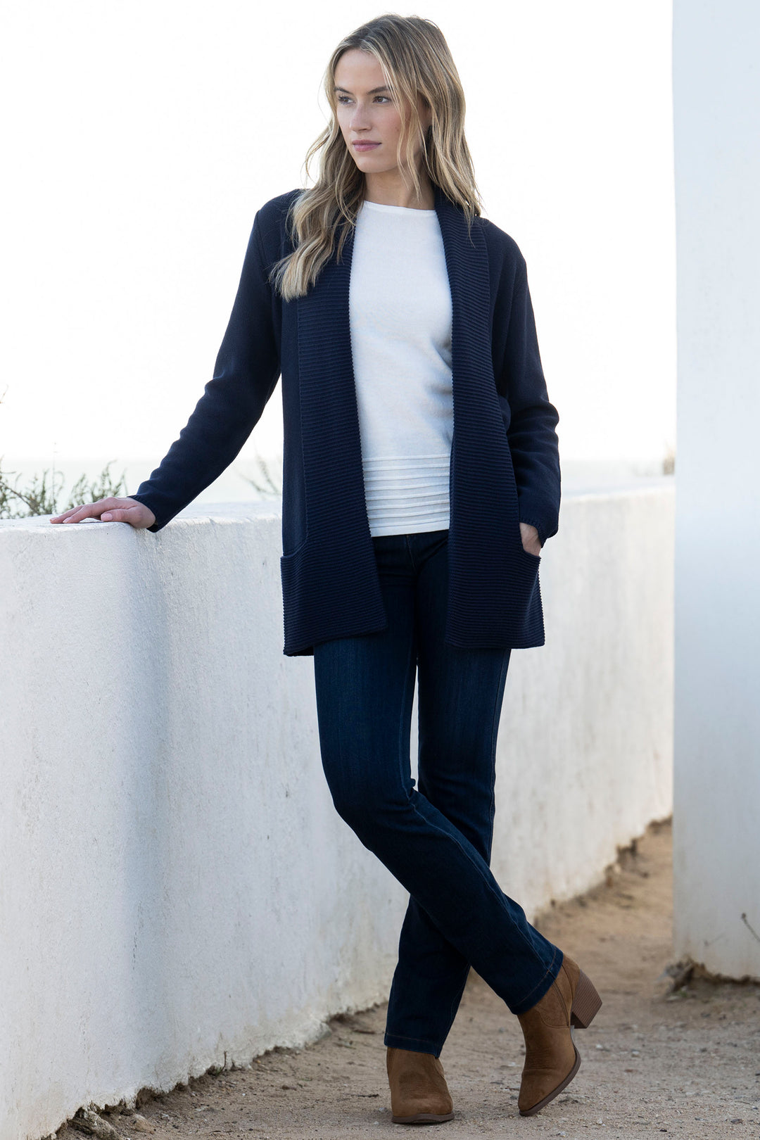 Woman wearing a navy cardigan, white shirt, and dark jeans standing against a white wall.