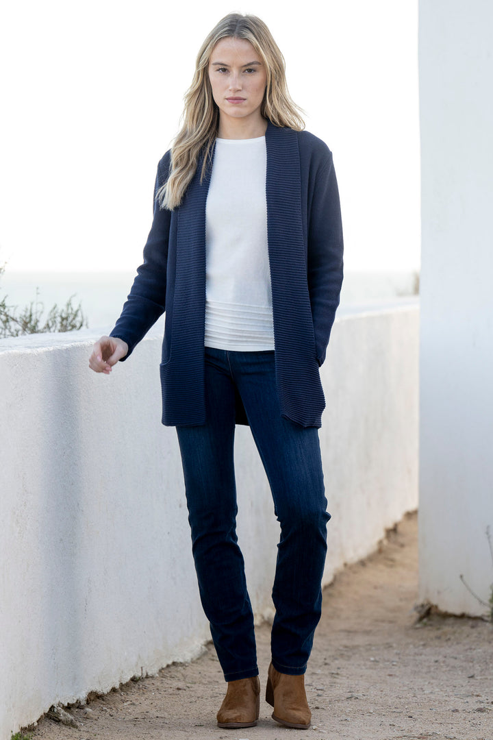 Woman wearing a navy cardigan, white shirt, dark jeans, and brown boots against a white wall.