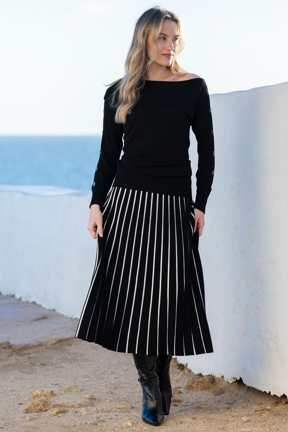 Woman wearing a black off-shoulder top and black and white striped skirt standing against a white wall.
