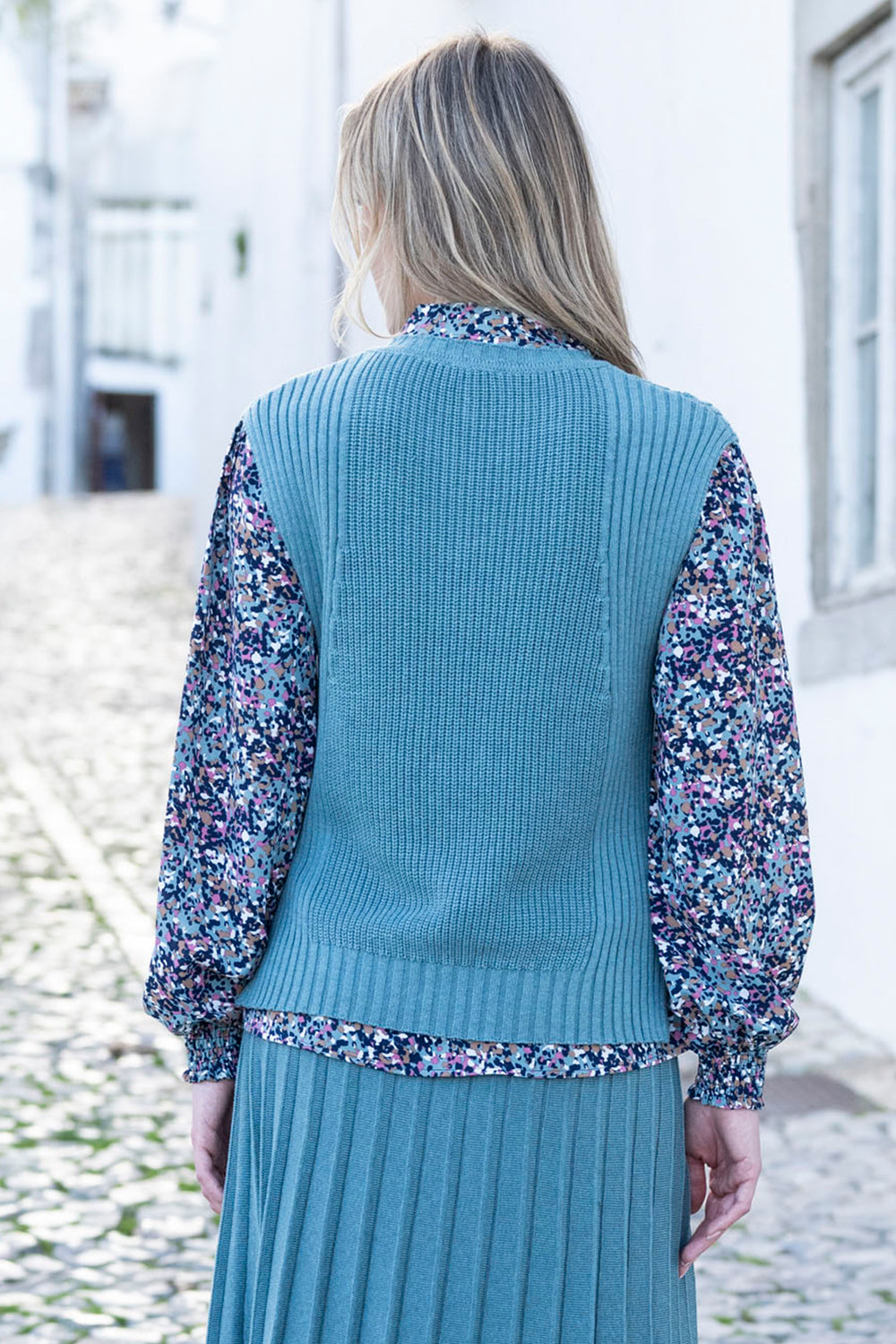 Person wearing a blue knitted vest over a floral blouse and skirt.