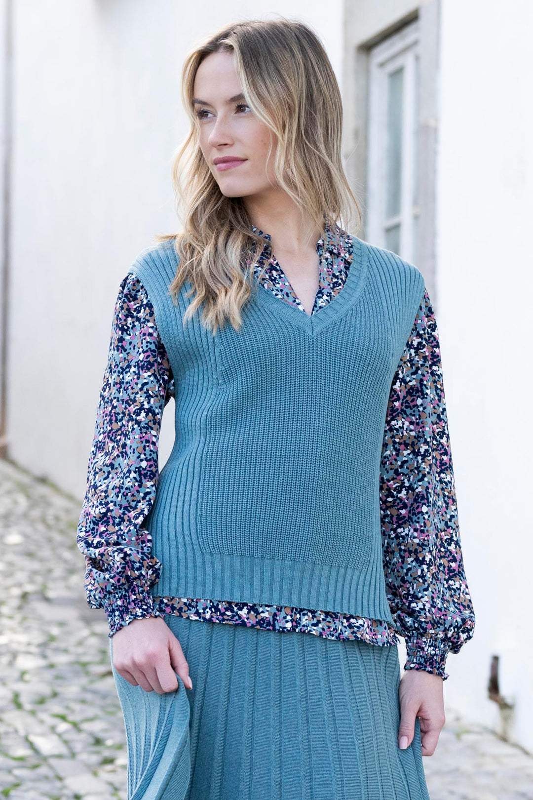 Woman wearing a teal ribbed sweater over a floral blouse with a long skirt outdoors.