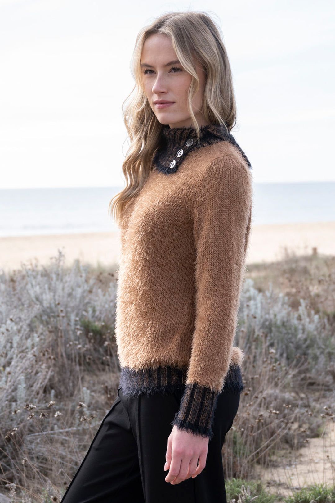 Woman wearing a brown sweater with black cuffs and collar standing on a beach.